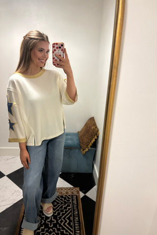 Woman taking a mirror selfie wearing a white sweater with colorful patterns and blue jeans.