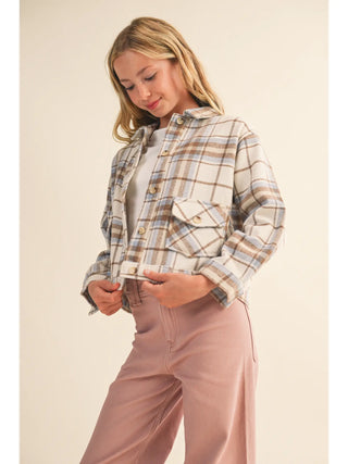 A woman wearing a cropped, long-sleeve plaid flannel shirt jacket in beige, brown, and blue tones, paired with dusty rose wide-leg pants, set against a solid light beige background.