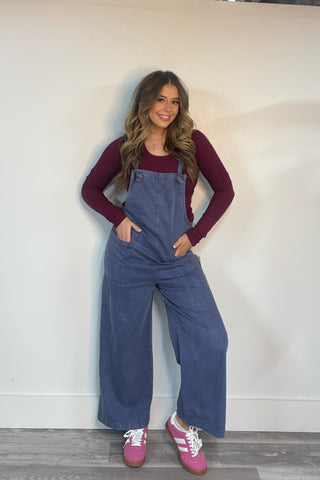 Woman wearing blue overalls and a maroon long-sleeve shirt against a white wall.