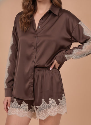 A woman wearing a brown satin pajama set with sheer lace sleeves and lace hem trim, photographed against a plain beige studio background.