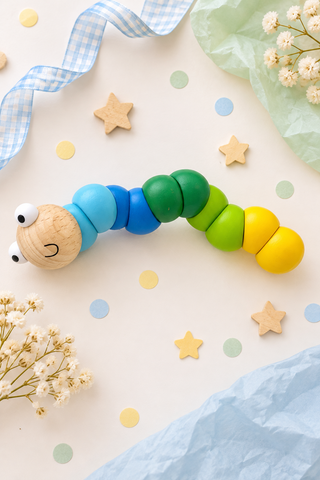 A wooden caterpillar toy with a natural wood head and segments painted light blue, medium blue, green, yellow, and orange, styled on a light cream background with paper stars, dots, ribbon, and dried flowers.