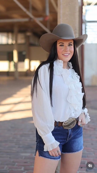 Woman wearing a white blouse with ruffles, denim shorts, and a brown cowboy hat outdoors.
