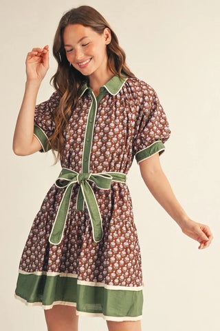 A woman modeling a short-sleeve collared dress with an allover brown and cream floral print, accented by solid olive green trim on the collar, placket, cuffs, and waist tie belt.