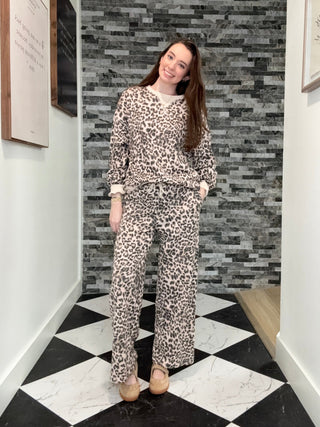 A woman stands in a hallway wearing a matching beige and black leopard-print lounge set consisting of a long-sleeve top and wide-leg pants, paired with tan flat shoes.