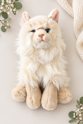 A fluffy cream-colored plush cat toy with blue eyes, pink nose, and tan paws, photographed on a light beige background with decorative greenery and a knitted textile in the corners.