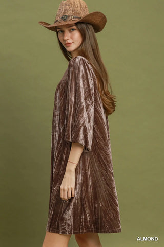 A woman wearing a brown velvet short dress with elbow-length sleeves and a matching cowboy hat, photographed against a solid olive green studio background. The word 'ALMOND' appears in the bottom right corner.