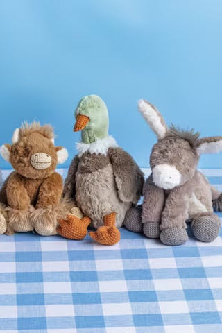 Five plush farm animal toys arranged in a row on a blue-and-white checkered surface against a light blue background. The toys include a white sheep, a brown chicken, a brown cow, a green duck, and a gray donkey.