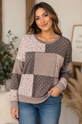 A woman wearing a long-sleeve patchwork sweatshirt with brown, beige, and white patterns, paired with blue jeans, in a cozy indoor setting.