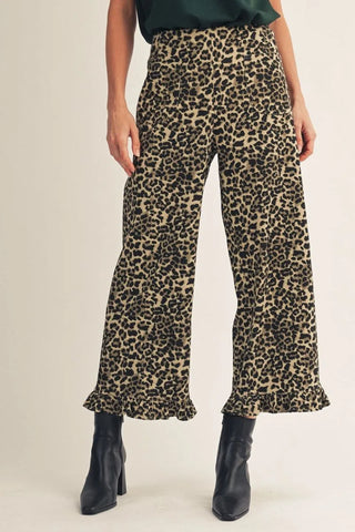 A model wearing high-waisted leopard print wide-leg pants with a ruffled hem, paired with black ankle boots, shown from the waist down against a plain light beige studio background.
