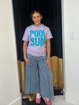 Person wearing a 'POOL BUM' t-shirt and wide-leg jeans standing in a doorway.