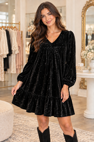 A woman models a black velvet long-sleeve mini dress with small silver rhinestone accents, paired with black cowboy boots, inside a boutique setting.