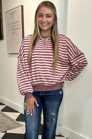 A woman modeling a pink and white striped long-sleeve sweatshirt with a crew neck, paired with distressed blue jeans and white sneakers, standing in an indoor hallway with a black and white checkered floor.