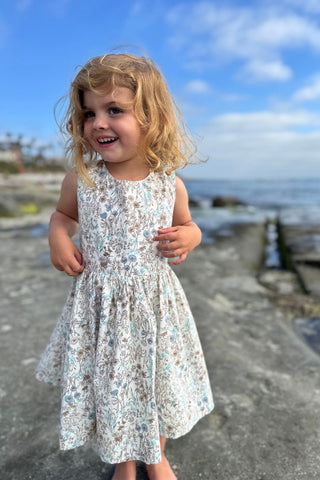 Young girl in a floral dress standing on a pathway near water with a blue sky.