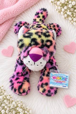 A colorful plush leopard toy with a pink, yellow, and black spotted pattern, lying on a white faux-fur surface with pink felt hearts, white baby's breath flowers, and a pink blanket. A hangtag reading Webkinz is attached to the toy.