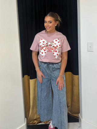 Person wearing a pink t-shirt with a heart and bow design, standing in front of a black and gold curtain.