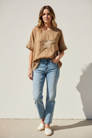 Woman wearing a brown t-shirt with a star design and light blue jeans against a white wall.