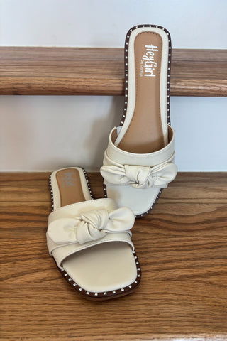 Pair of cream-colored sandals with bow details on a wooden surface.