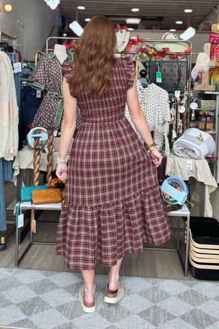 A woman viewed from behind wearing a red and brown plaid midi dress with ruffled sleeves and a tiered hem, standing inside a clothing store.