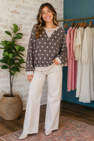 A woman modeling a brown long-sleeve blouse with white polka dots and cream wide-leg pants in a boutique setting.