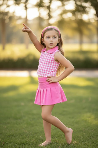 Young girl in a pink checkered top and skirt standing on grass, pointing upwards.