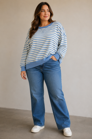 A woman modeling a light blue and white striped crewneck sweatshirt with blue jeans and white sneakers, standing against a neutral beige studio background.