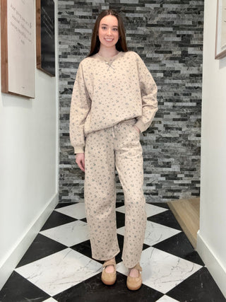A woman stands in a hallway wearing a matching beige pajama set with a small floral print, consisting of a long-sleeve top and wide-leg pants, paired with tan flats.