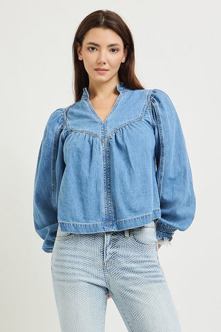 A woman wearing a light blue denim cropped jacket with puffed sleeves and a gathered neckline, paired with light-wash jeans, against a plain light gray studio background.