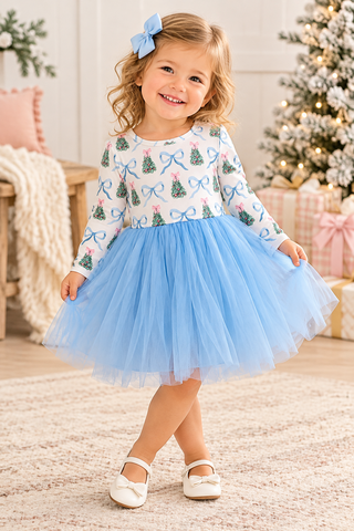 A young girl with blonde curly hair wearing a white long-sleeve dress with a blue bow and tree pattern on the bodice and a light blue tulle skirt, standing in a cozy holiday room with a Christmas tree in the background.