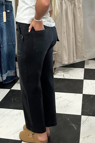 A woman modeling black wide-leg cropped pants in a clothing store, standing on a black and white checkered floor with racks of clothes in the background.
