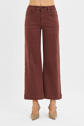 A person wearing rust-brown wide-leg cropped jeans with beige pointed-toe heels, shown from the waist down against a plain light gray studio background.