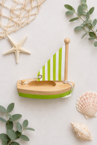 A wooden toy sailboat with a green and white striped sail and a green hull trim, styled on a light background with seashells, eucalyptus leaves, a starfish, and a net.