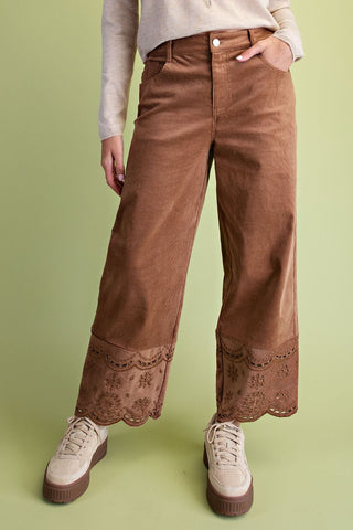 A person wearing brown wide-leg corduroy pants with a scalloped lace hem, styled with a cream long-sleeve top and beige platform sneakers against a light green studio background.