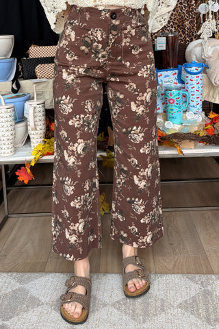 A person wearing brown wide-leg cropped pants with a beige and white floral print, paired with brown slide sandals, standing in a retail store setting.