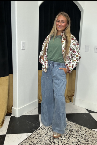 A woman stands indoors wearing a green crewneck top, light blue wide-leg jeans, and a floral-patterned bomber jacket. She is posing in front of a white wall with black and gold curtains and a black-and-white checkered floor.