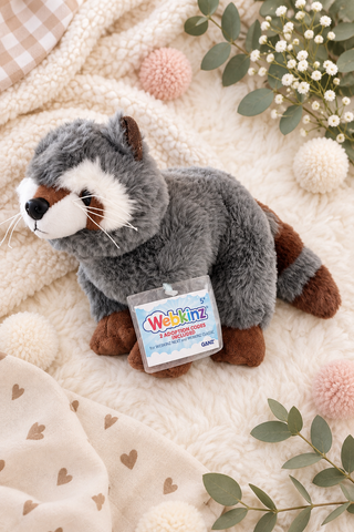 A plush raccoon toy by Webkinz, styled on a cozy cream-colored blanket with decorative pom-poms, eucalyptus leaves, and a checkered throw. The raccoon is gray with white and brown details, and a product tag is attached to its neck.