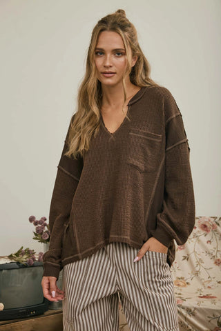 A woman wearing a brown ribbed knit long-sleeve top with a V-neck and striped pants, standing in a softly lit indoor setting.