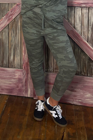 Person wearing camouflage jogger pants and black and white sneakers against a wooden background