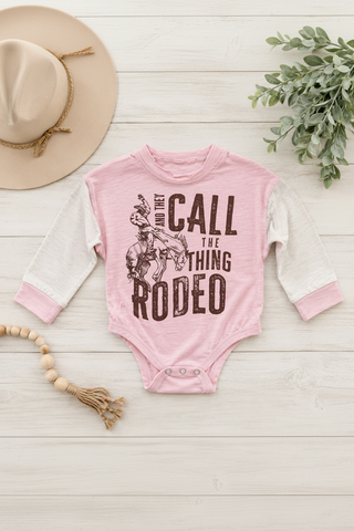 Pink baby onesie with 'And they call the thing rodeo' text on a wooden surface with a hat and plant.