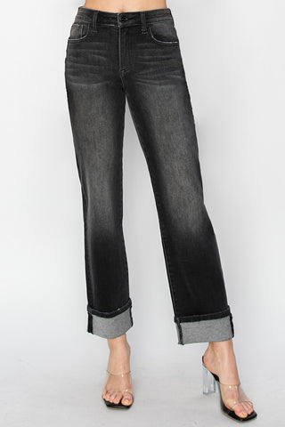 A person wearing dark gray to black faded straight-leg jeans with rolled cuffs, paired with clear high-heeled sandals, against a light gray studio background.