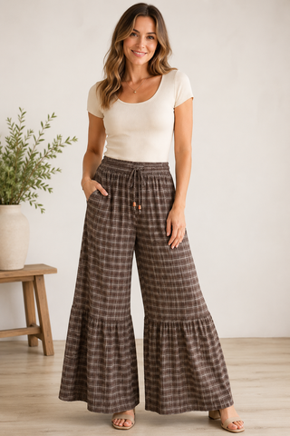 A woman modeling a pair of brown and beige plaid wide-leg ruffled pants with a drawstring waist, paired with a cream short-sleeve top, standing in a brightly lit indoor setting.