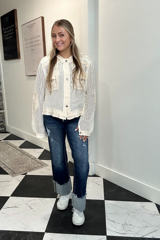 A woman stands in a hallway wearing a cream and white vertically striped button-up shirt with ruffled trim, blue jeans with cuffed hems, and white sneakers.