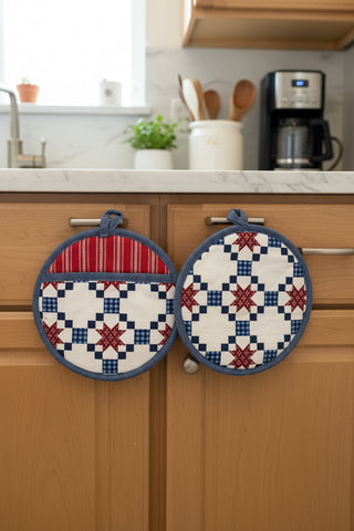 Pair of quilted pot holders with star pattern hanging on a wooden cabinet.