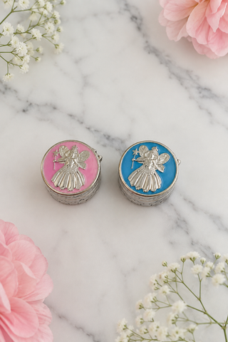 Two round silver-toned keepsake boxes with embossed angel figures, one with pink enamel and one with blue enamel, displayed on a white marble surface with pink flowers and baby's breath.