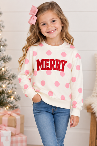 A young girl wearing a cream crewneck sweatshirt with pink polka dots and a red 'MERRY' graphic, paired with blue jeans and white sneakers, standing in a cozy holiday room next to a decorated Christmas tree.
