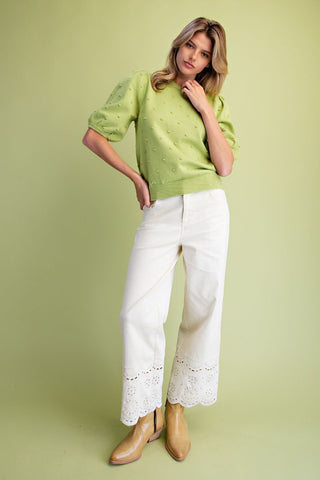 A woman modeling a light green short-sleeve knit sweater with textured knit details, paired with white wide-leg pants featuring lace trim at the hem and tan ankle boots, set against a solid light green studio background.