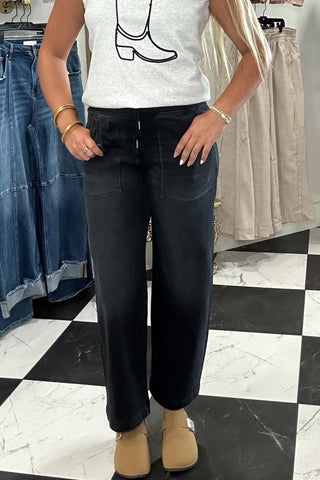 A woman stands in a clothing boutique wearing a white sleeveless top with a black cowboy boot graphic, paired with dark wide-leg jeans and tan boots. The background shows racks of other clothing and a black-and-white checkered floor.