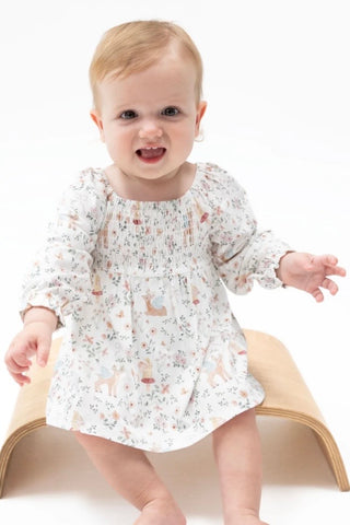 A baby girl wearing a long-sleeve smocked dress with a woodland deer print, sitting on a wooden arch stool against a white background.