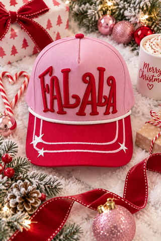 A pink and red baseball cap with 'FALALA' embroidered in red across the front, surrounded by Christmas decorations including a wrapped gift, candy canes, pinecones, ornaments, and a mug of hot chocolate.
