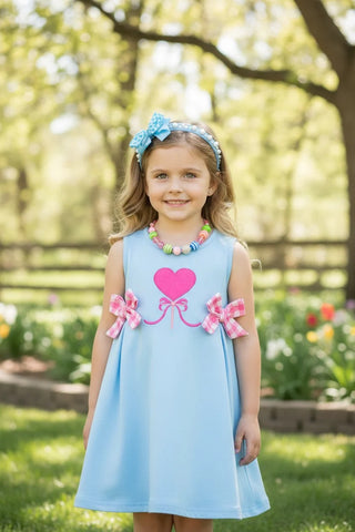 Young girl in a light blue dress with pink heart and bow details, standing outdoors.