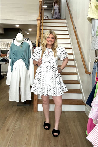 Woman in a polka dot dress standing in a store with clothing racks and stairs in the background.